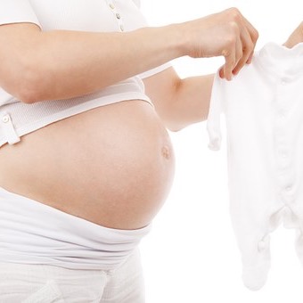 Auf dem Bild sieht man einen Schwangerschaftsbauch. Die Schwangere hält einen weißen Babystrampler vor den Bauch.- Ernährung in der Schwangerschaft ist ein ganz wichtiges Thema.